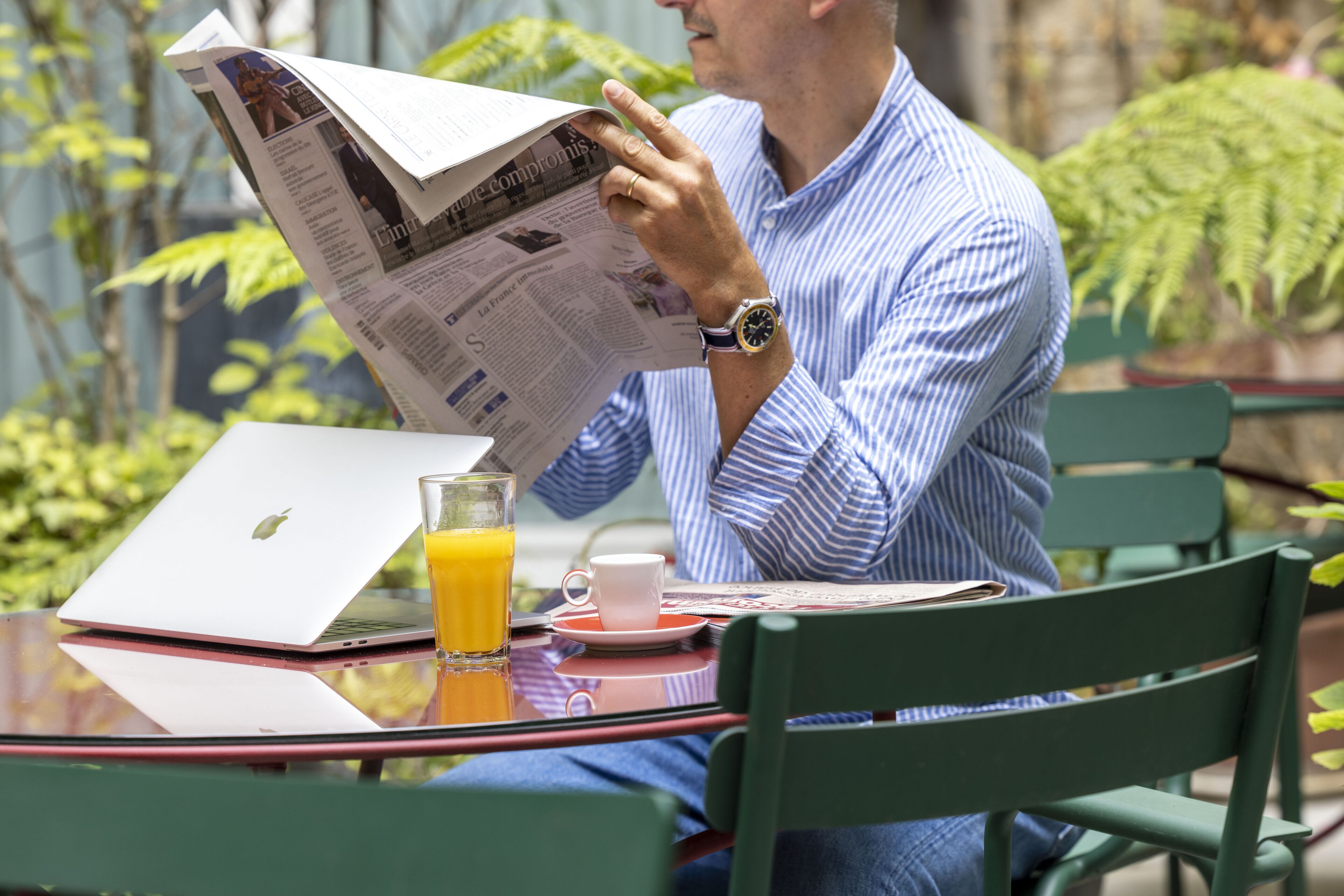 Iemand leest een krant aan een tafel, met een laptop en een glas sinaasappelsap.