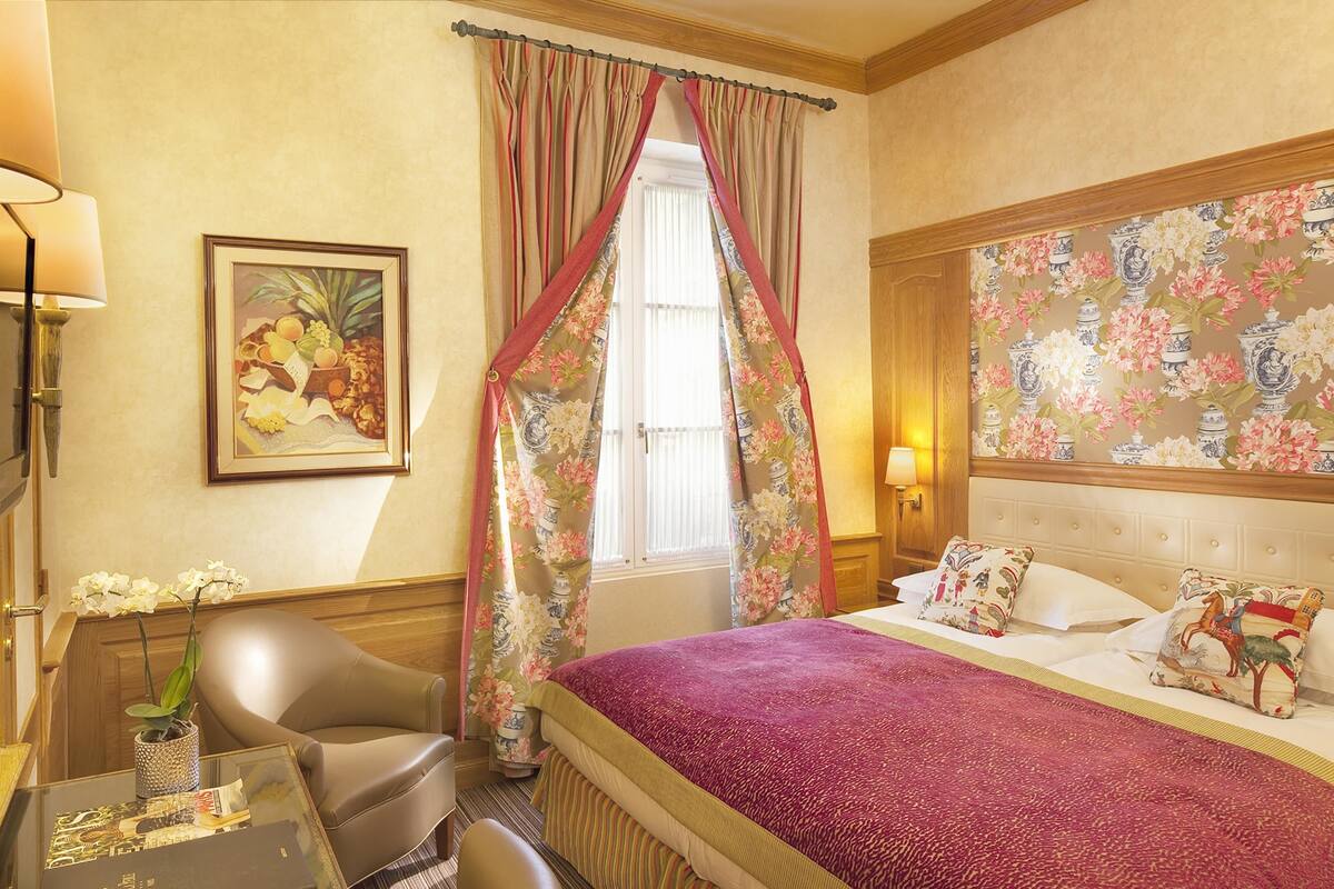 A hotel room with a bed, a chair, a desk, a lamp, and a window with curtains.