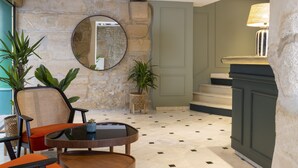 A lobby with a stone wall, a round mirror, a staircase, and a table with chairs. - Hotel La Perle (Paris)