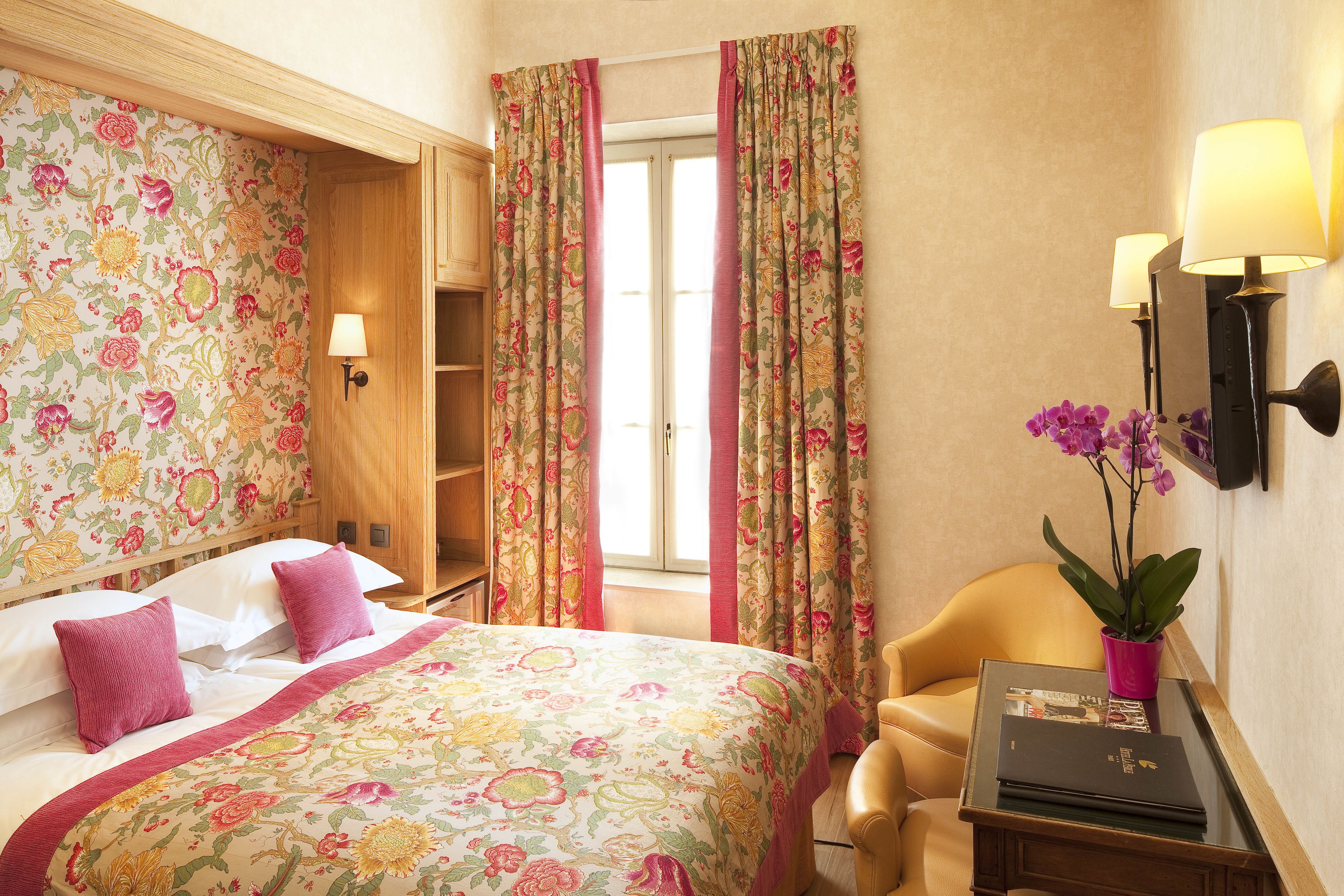 A hotel room with a floral bedspread, a wooden wardrobe, a desk with a lamp, and a chair.