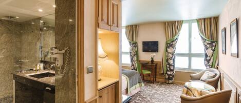A hotel room with a marble bathroom, a bed with a floral bedspread, a small table, a chair, and a TV on the wall.