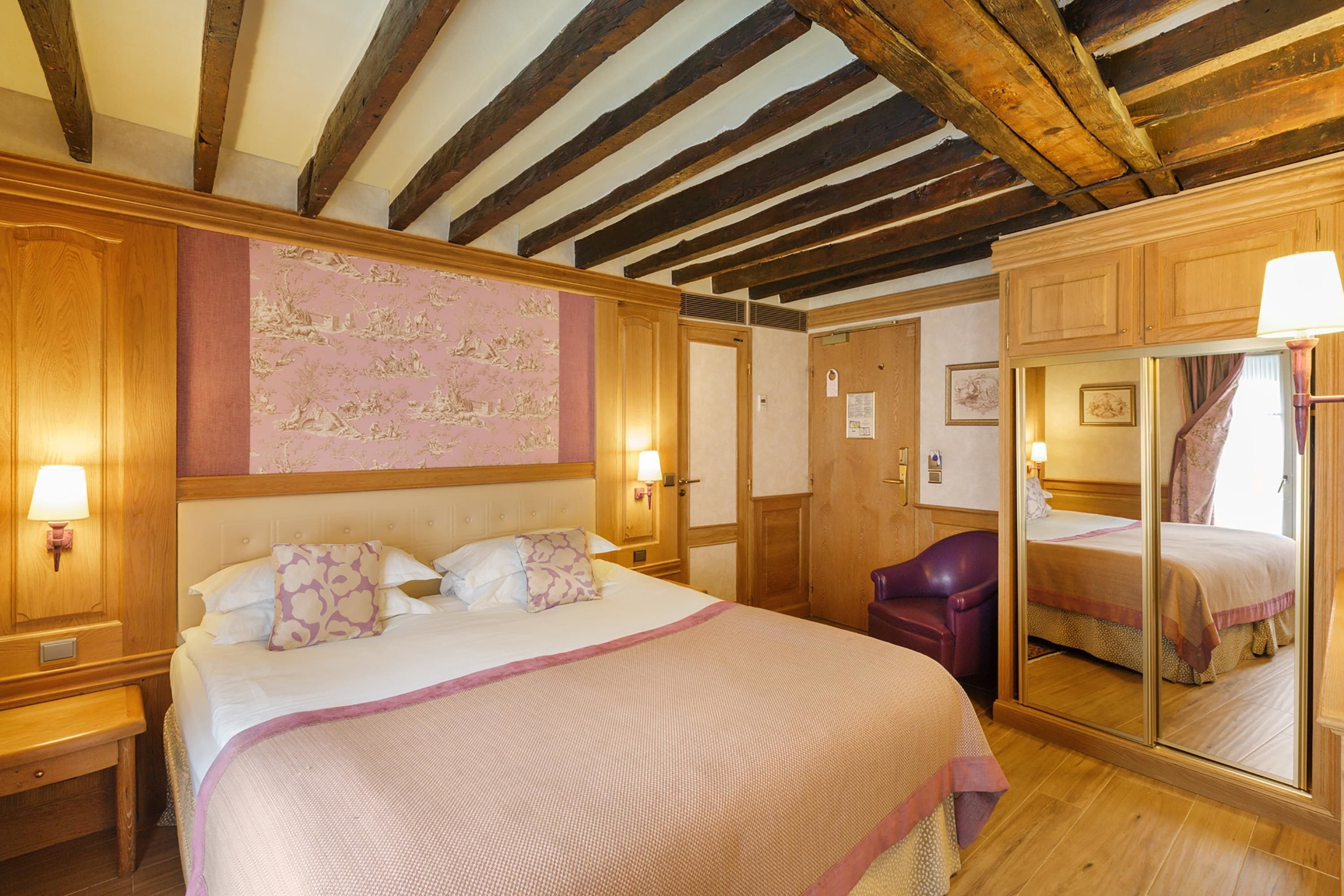 A hotel room with a large bed, wooden ceiling beams, a mirror, and a purple chair.