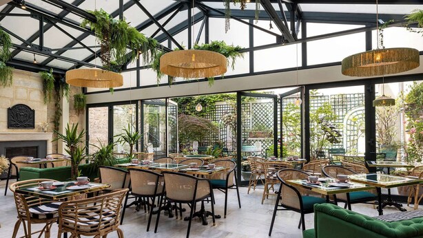 An indoor dining area with wicker chairs and tables, a glass ceiling, and greenery. - Hotel La Perle (Paris)