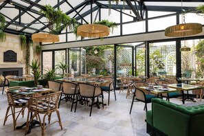 An indoor dining area with wicker chairs and tables, a glass ceiling, and greenery. - Hotel La Perle (Paris)