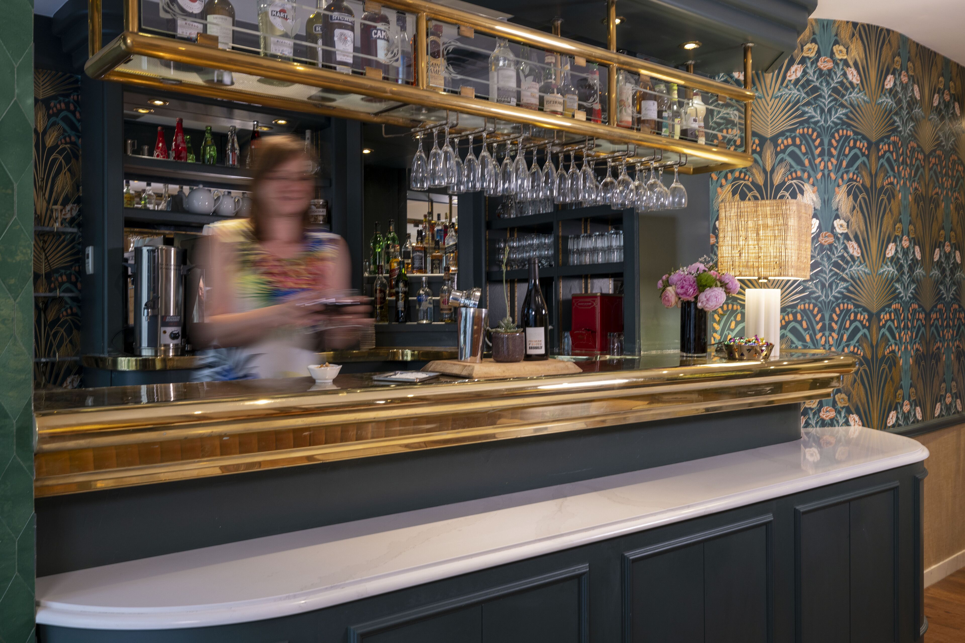 Un espace bar avec des étagères remplies de bouteilles et de verres, un comptoir agrémenté d’un vase de fleurs et un mur orné de motifs.