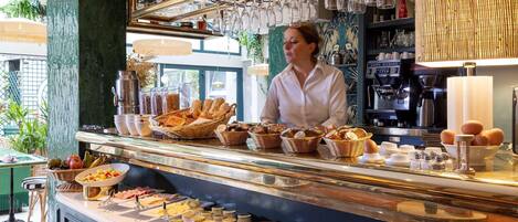 Un bufé de desayuno con diversos alimentos, como pan, bollería y huevos, en una sala luminosa que cuenta con una máquina de café y copas de vino a la vista.