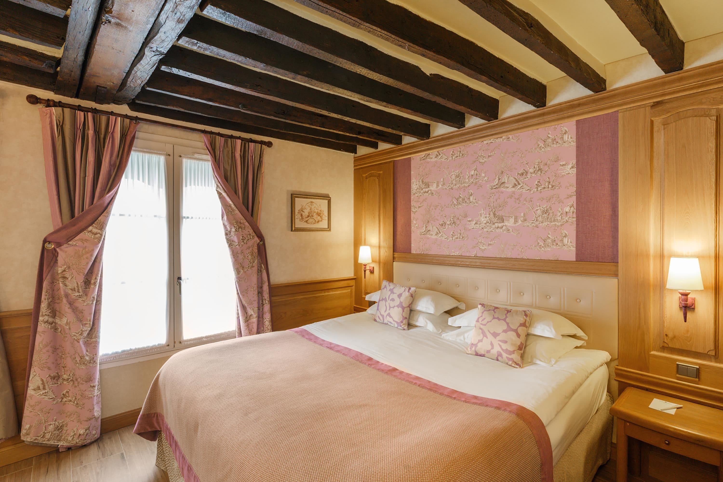 A bedroom with a large bed, wooden beams on the ceiling, a window with curtains, and a wall-mounted lamp.