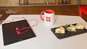 A modern room with a wooden bookshelf, a flat-screen TV displaying 'ibis hotels', a tablet, a mug, and a plate of food. - Hotel ibis El Jadida (El Jadida)