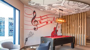 A pool table in a modern room with a large window, a blue chair, and a wall mural with musical notes and a guitar. - Hotel ibis El Jadida (El Jadida)