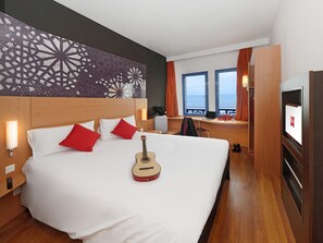 Una habitación de hotel con una cama grande, una guitarra, un escritorio con una computadora portátil y un televisor.