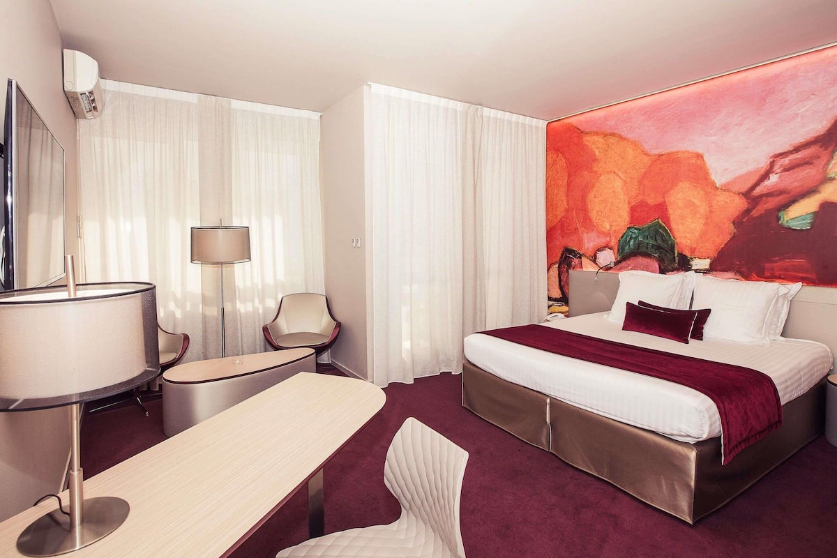 A hotel room with a bed, a desk, a chair, a lamp, and a large abstract painting.