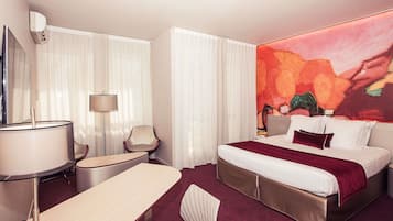 A hotel room with a bed, a desk, a chair, a lamp, and a large abstract painting.