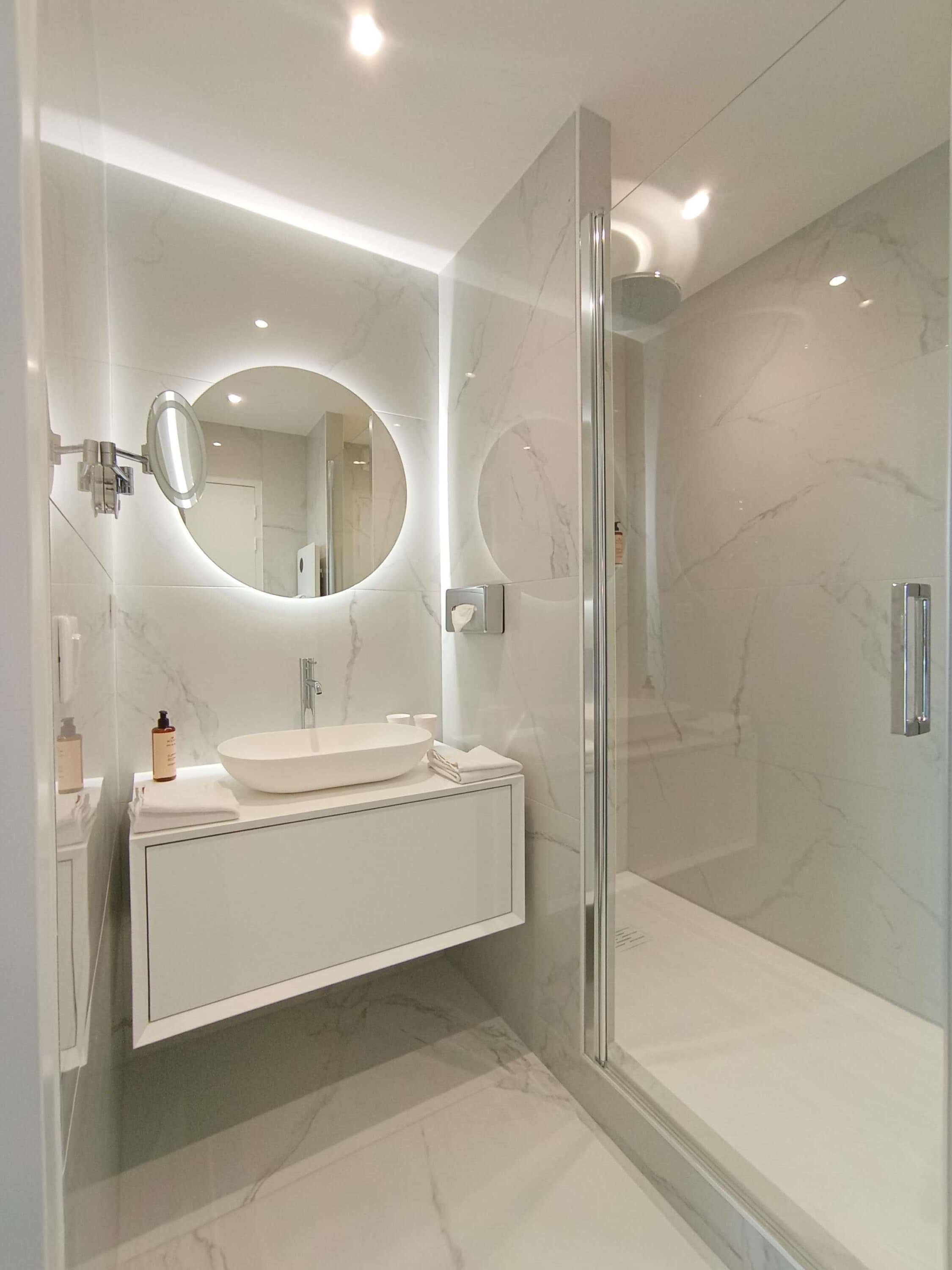 Un baño moderno con cabina de ducha de vidrio, un espejo redondo y un tocador blanco.