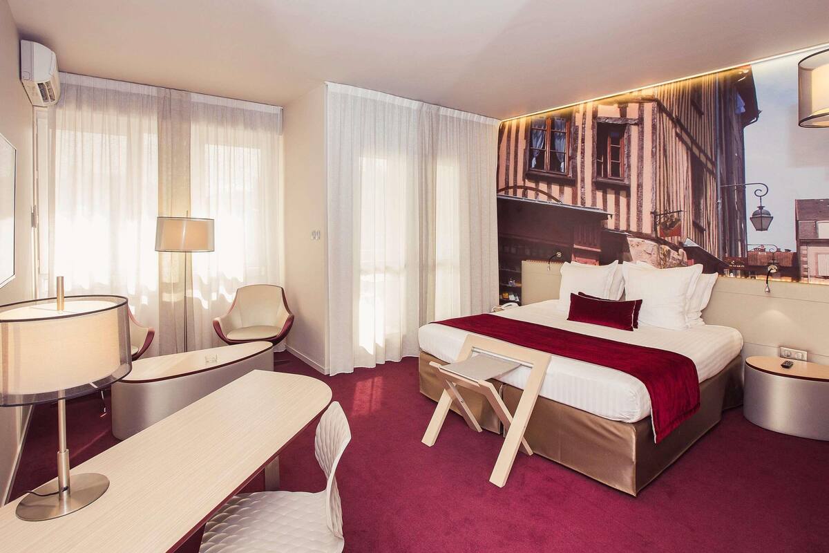 A hotel room with a bed, a desk, a chair, a lamp, and a large window with sheer curtains.