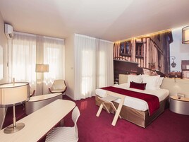 Een hotelkamer met een bed, een bureau, een stoel, een lamp en een groot raam met dunne gordijnen.