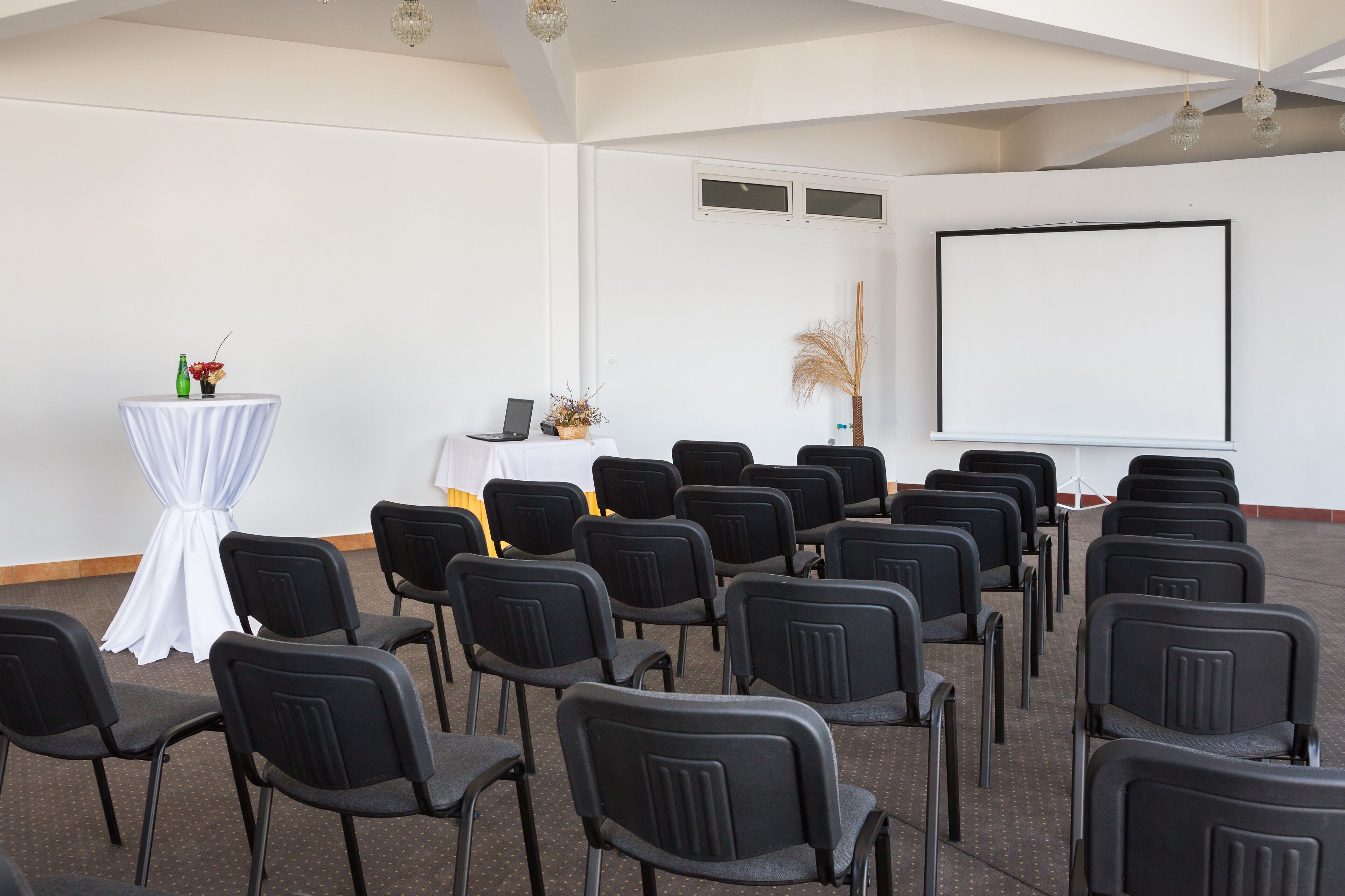 Un salón de conferencias con filas de sillas negras, una mesa blanca cubierta con un mantel, una pantalla de proyector y una pequeña mesa con una computadora portátil y adornos.