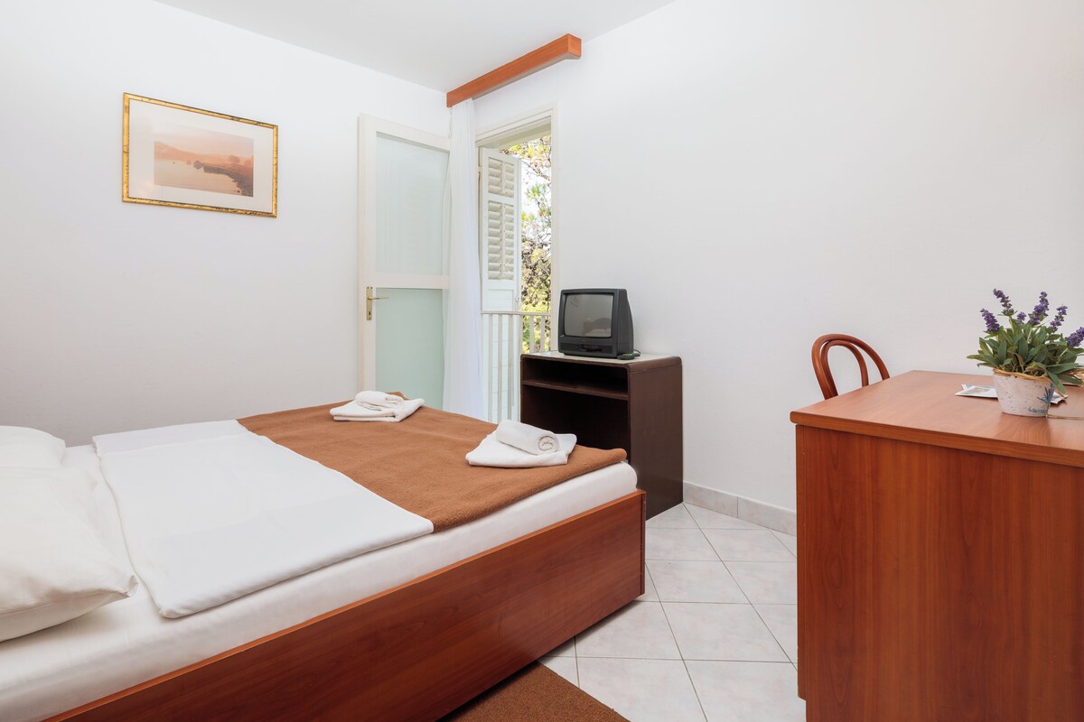 A bedroom with a bed, a desk, a television, and a window with blinds.