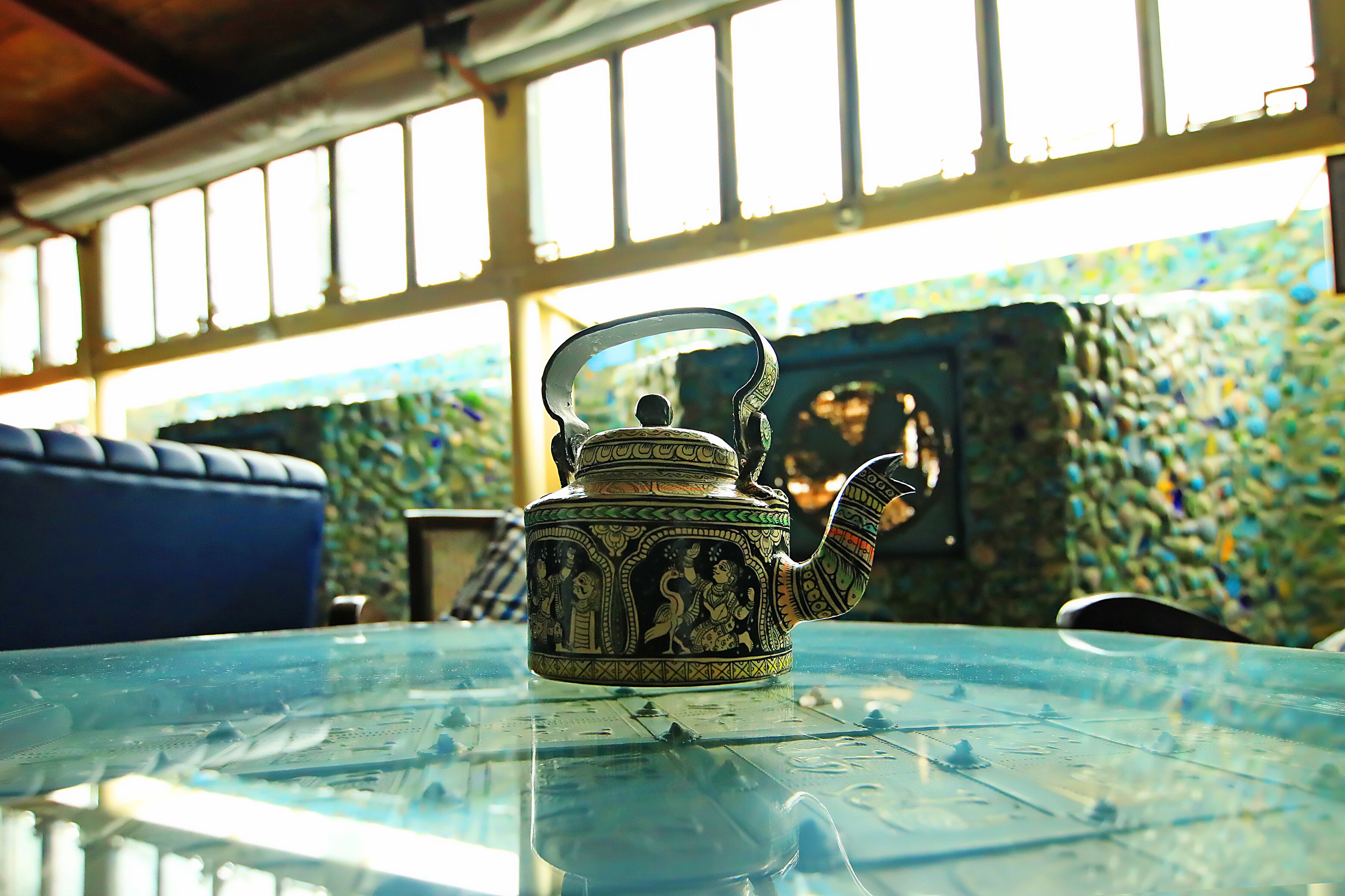 Una tetera sobre una mesa de vidrio, con vista a una pared de mosaicos y una silla azul al fondo.