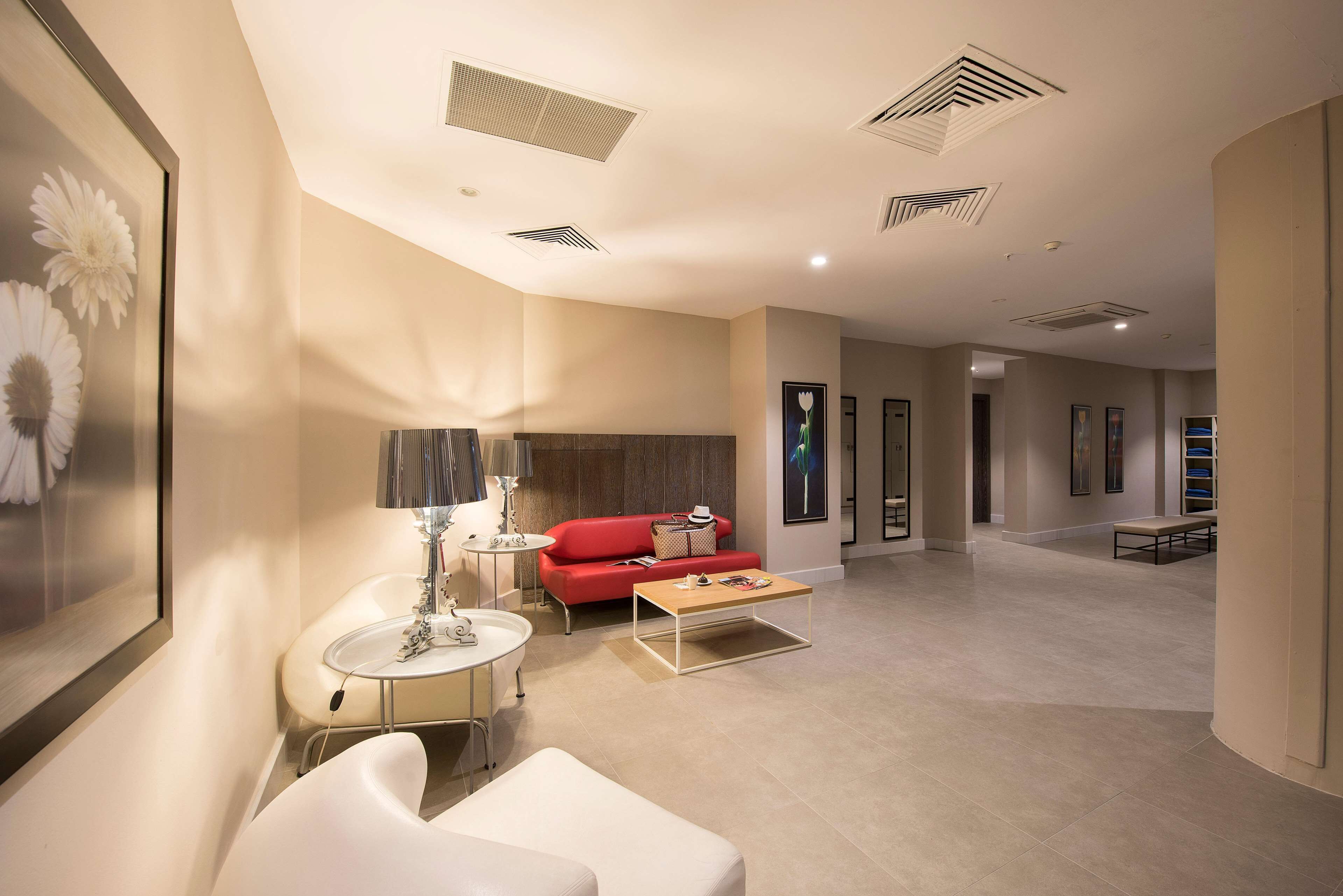 A spacious living area with a red sofa, a white armchair, a coffee table, and a lamp. There are ceiling vents and framed artwork on the walls.