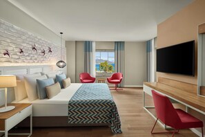 A modern hotel room with a large bed, a desk, two red chairs, a TV, and a view of the sea.