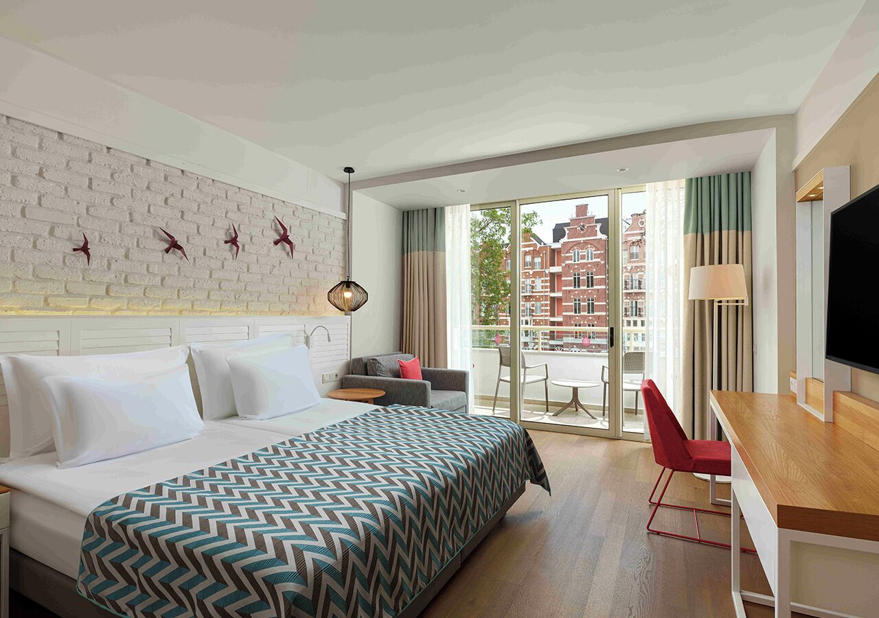A hotel room with a bed, a desk, a TV, and a balcony with a city view.