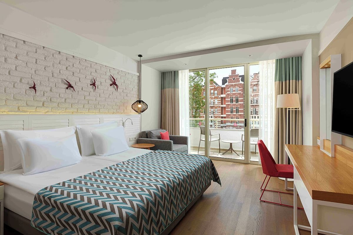 A hotel room with a bed, a desk, a TV, and a balcony with a city view.