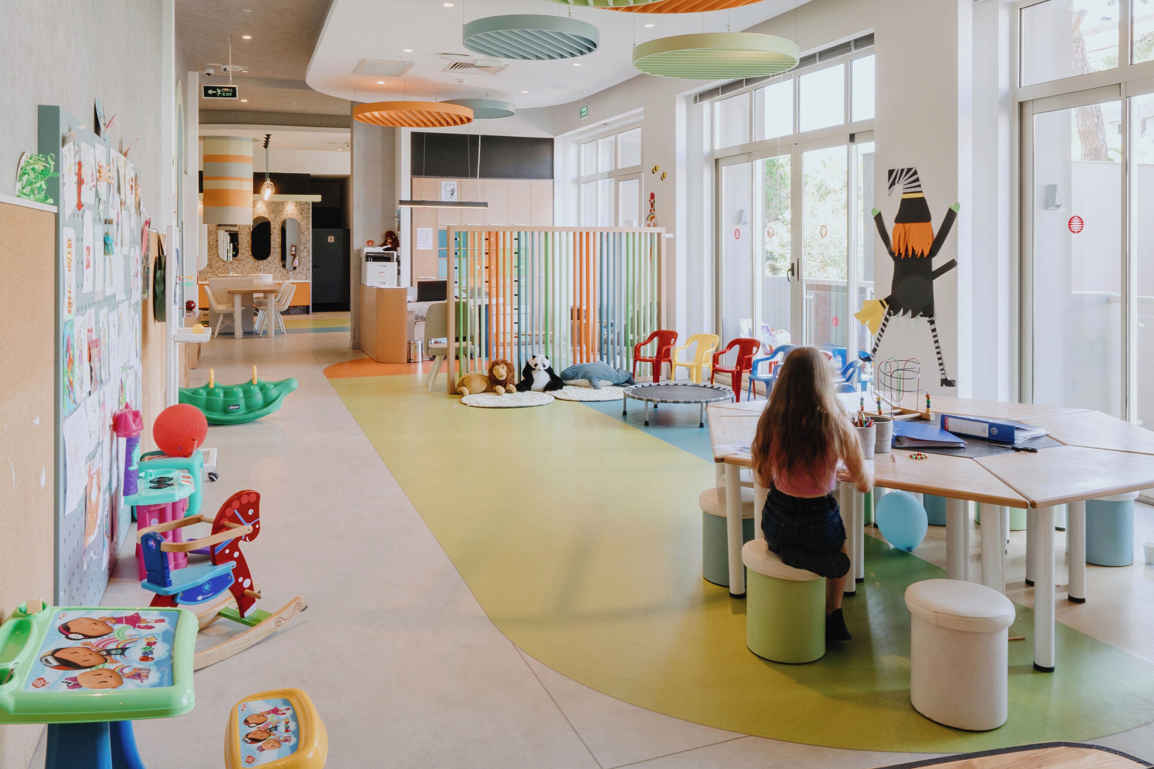 Ein Spielzimmer für Kinder mit bunten Möbeln, einem großen Fenster und einem Kind, das an einem Tisch spielt.