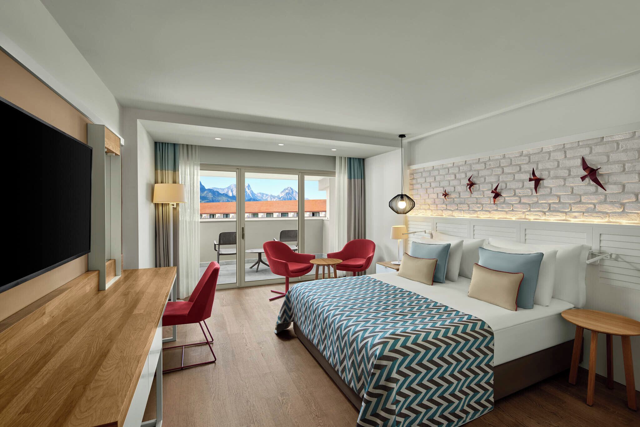 Habitación de hotel con cama, un escritorio, dos sillas rojas, una mesita pequeña, un televisor y vistas a la montaña desde la ventana.