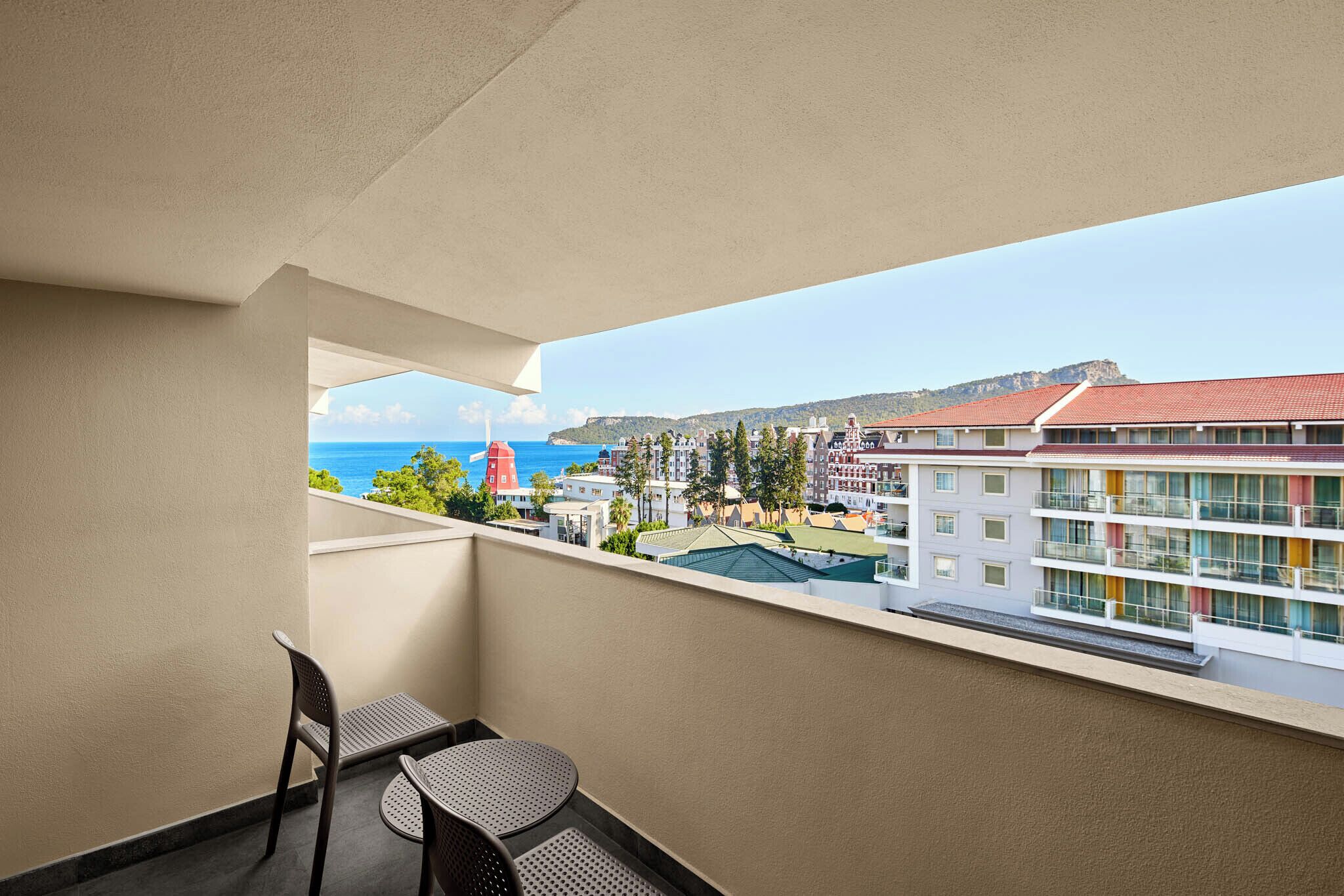 A balcony with a table and chairs, overlooking a coastal area with buildings and a red-roofed structure.