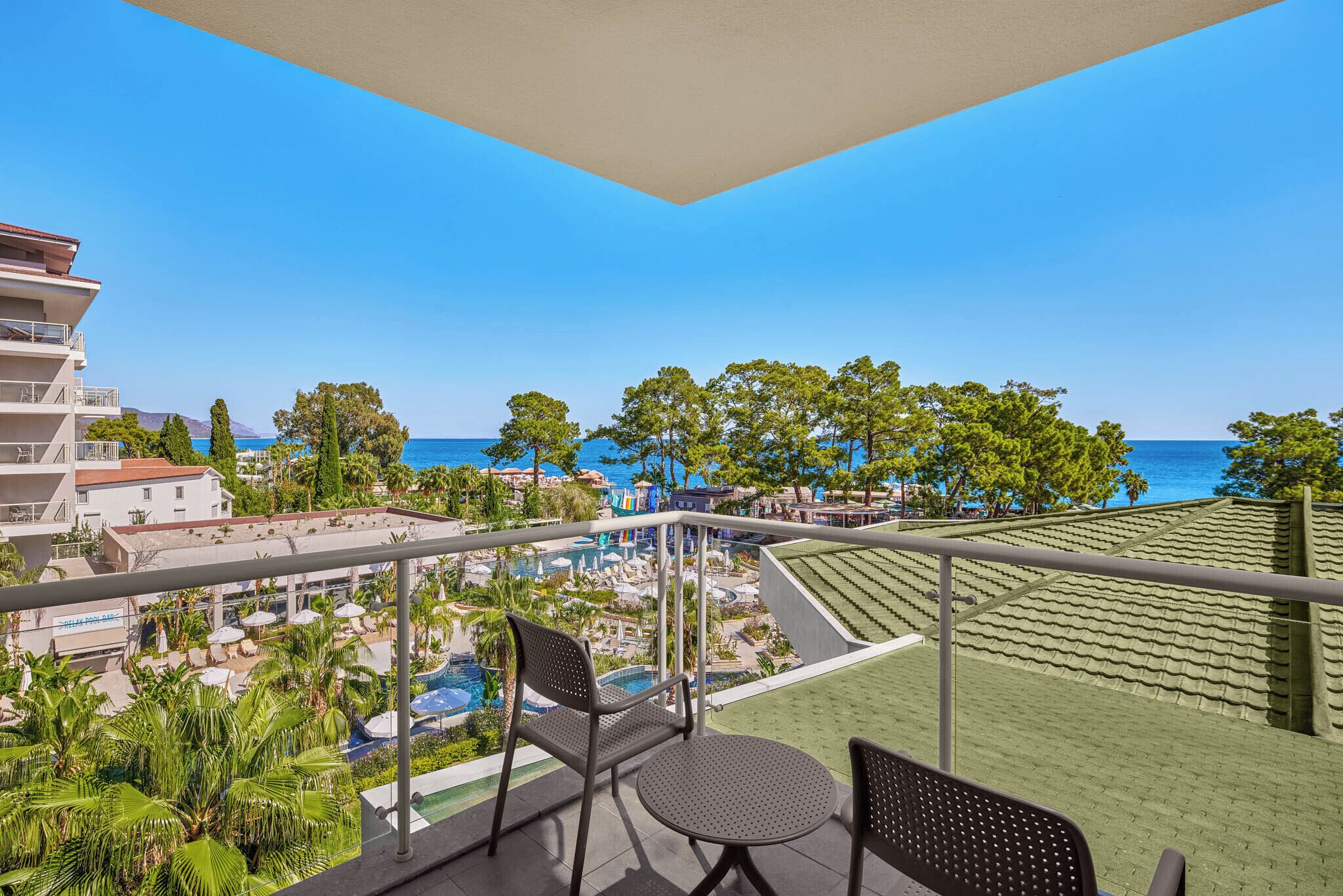 A balcony with a view of a pool and resort area, including a building with multiple floors and a clear blue sky.