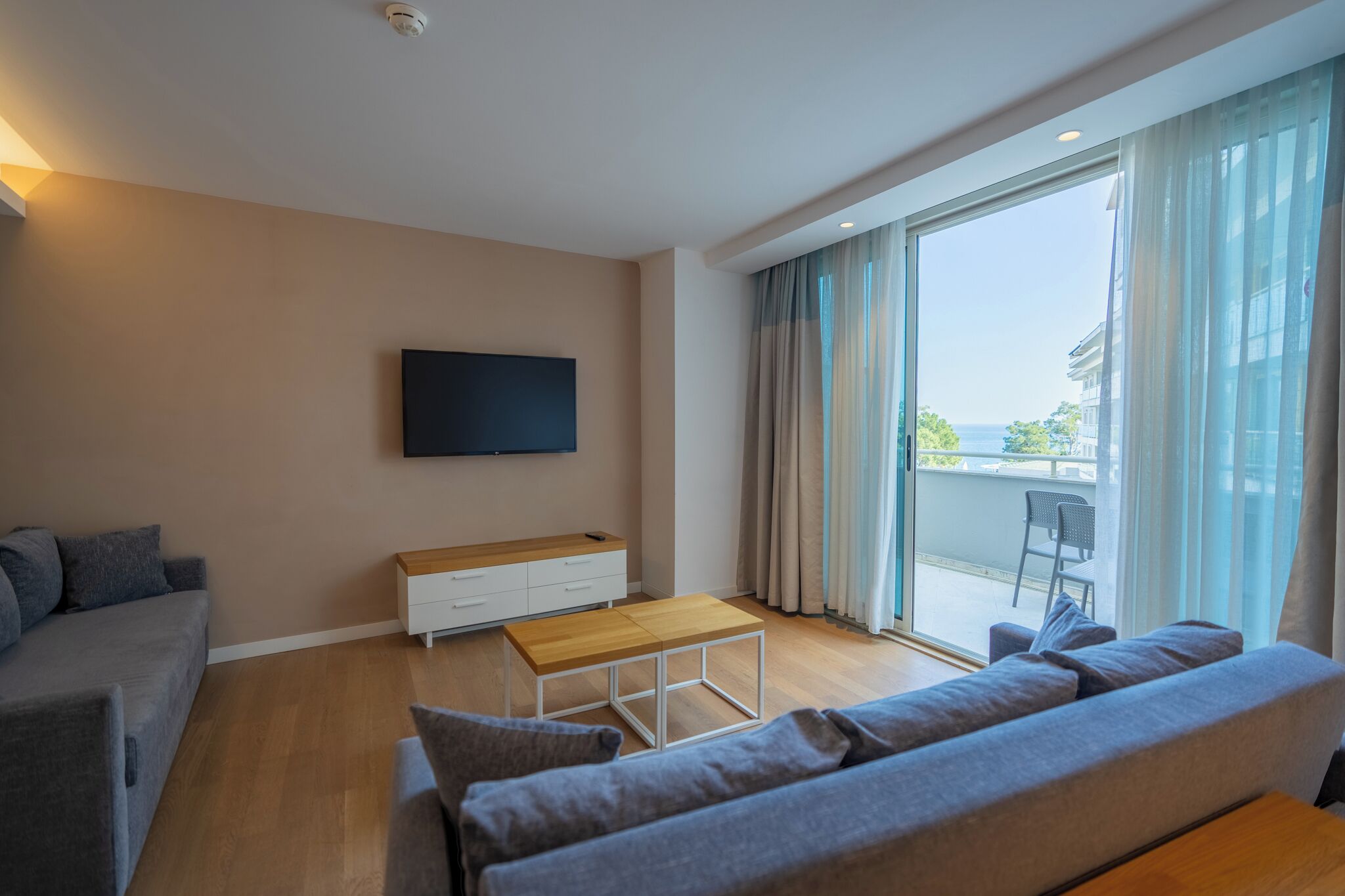 A modern living room with a sofa, a coffee table, a TV mounted on the wall, and a balcony with a view.