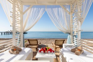 Un pabellón frente al mar con cortinas blancas, una mesa dispuesta con fruta y dos sillas.