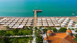 An aerial view of a beach resort with rows of sunbeds, a pier extending into the sea, and a central building with a dome roof. - Sherwood Exclusive Lara, All Inclusive (Antalya)