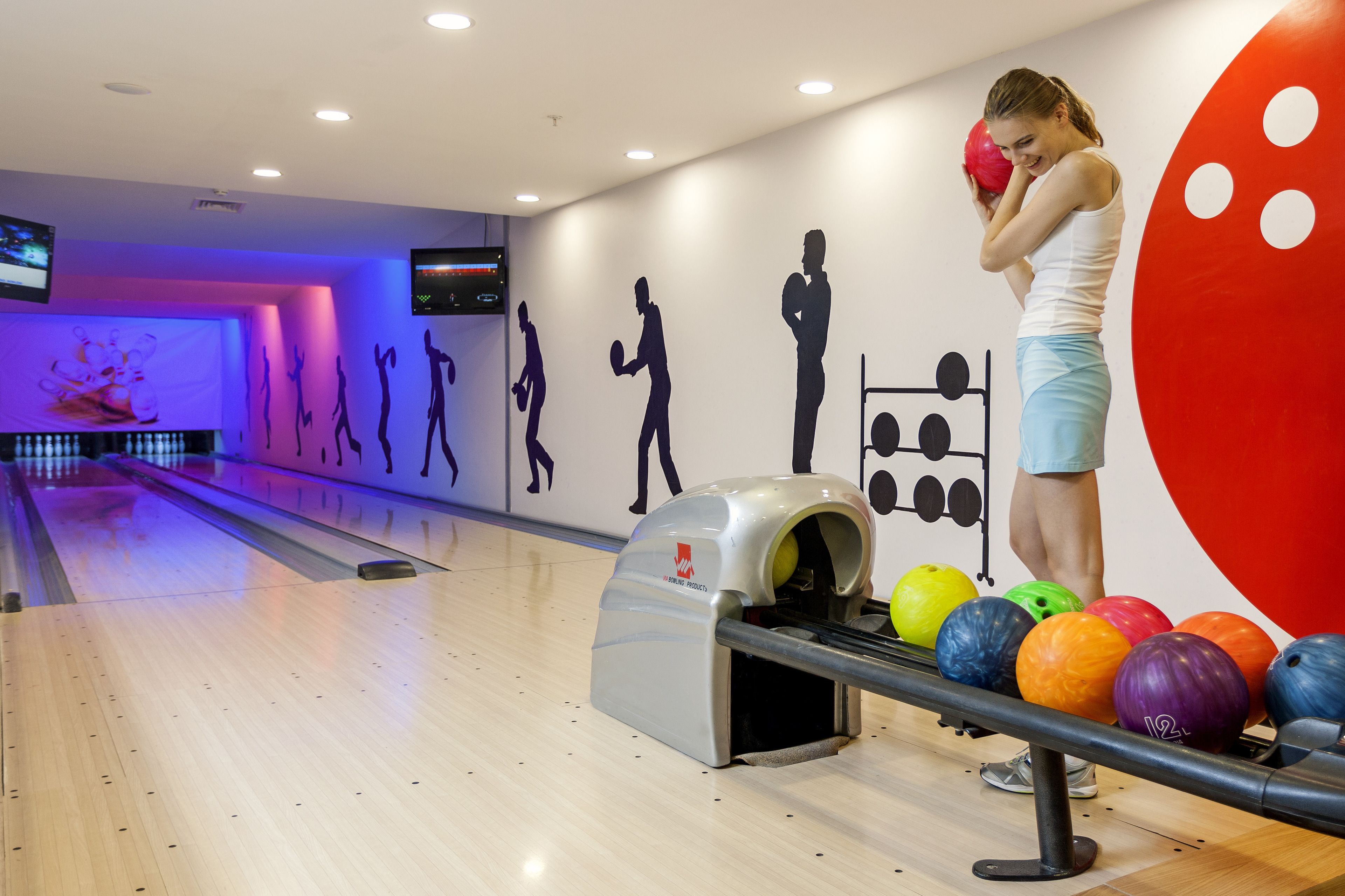 A bowling alley with a woman preparing to bowl, a bowling ball rack, and colorful bowling balls.