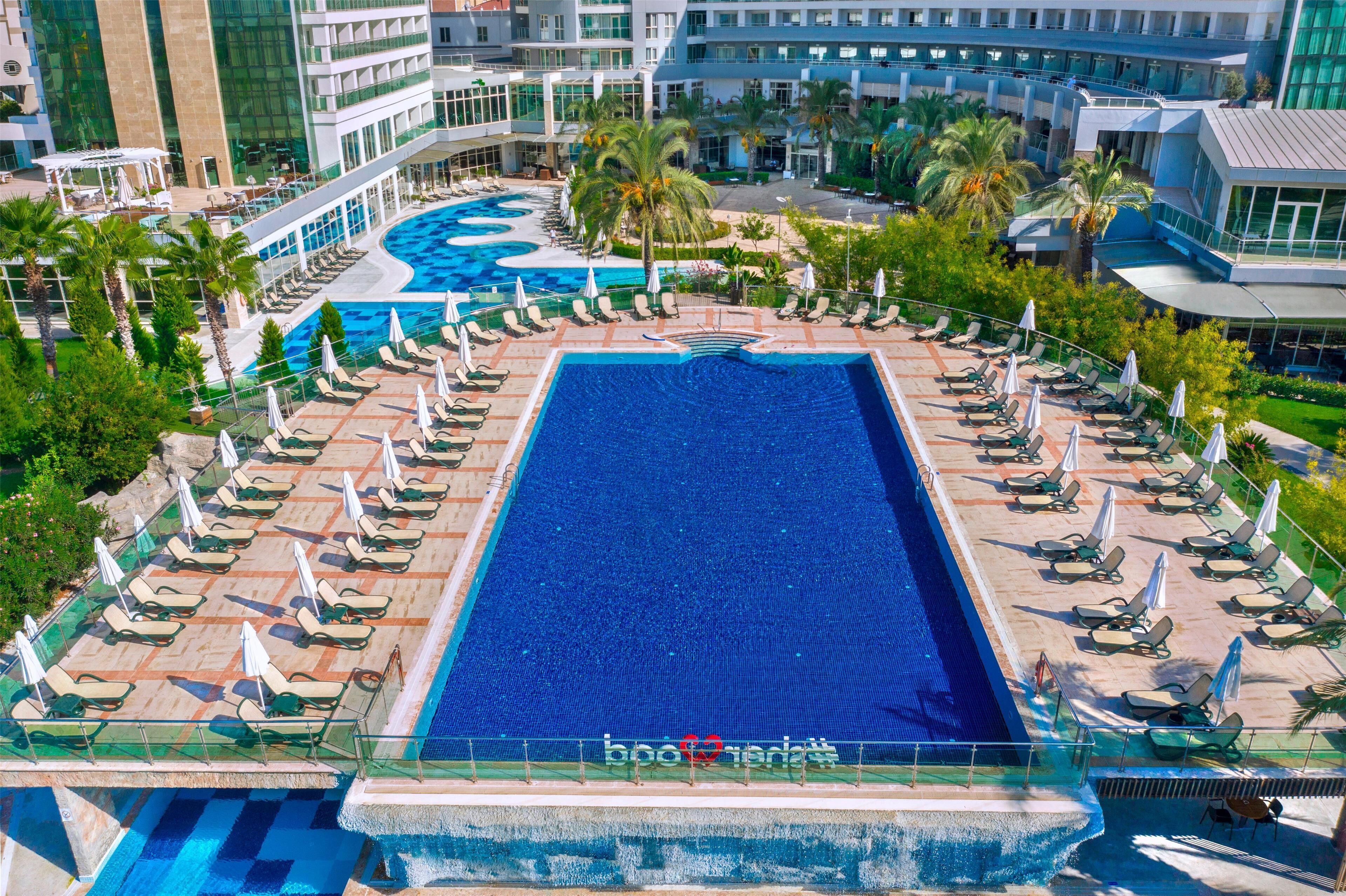 Une grande piscine rectangulaire entourée de chaises longues et de parasols, elle-même bordée d’une végétation luxuriante et de bâtiments modernes.