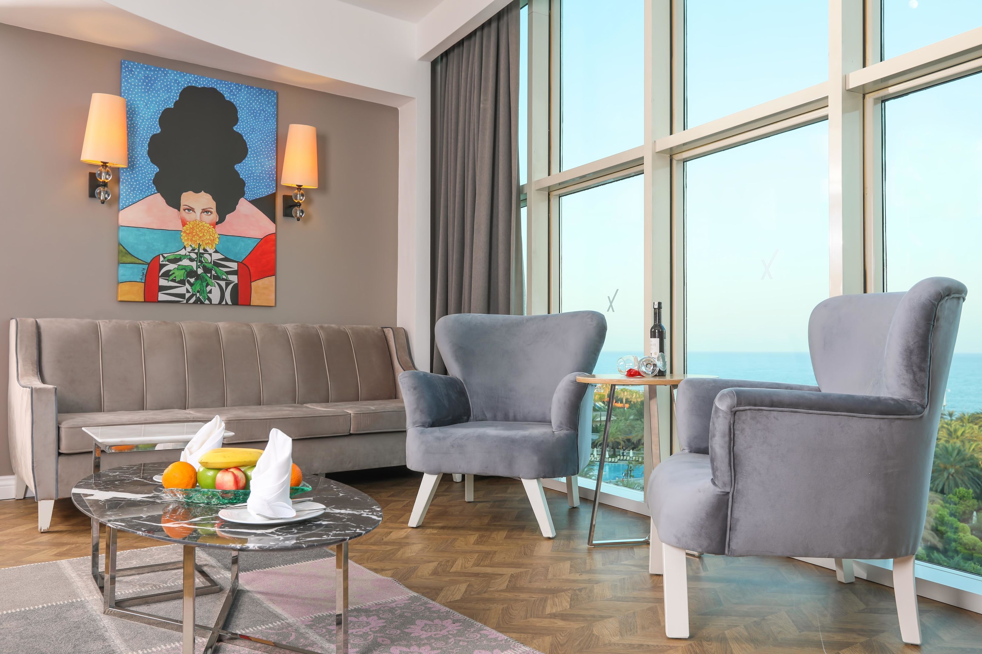 A modern room with a sofa, two armchairs, a small table with fruit and napkins, and a large window with a view.