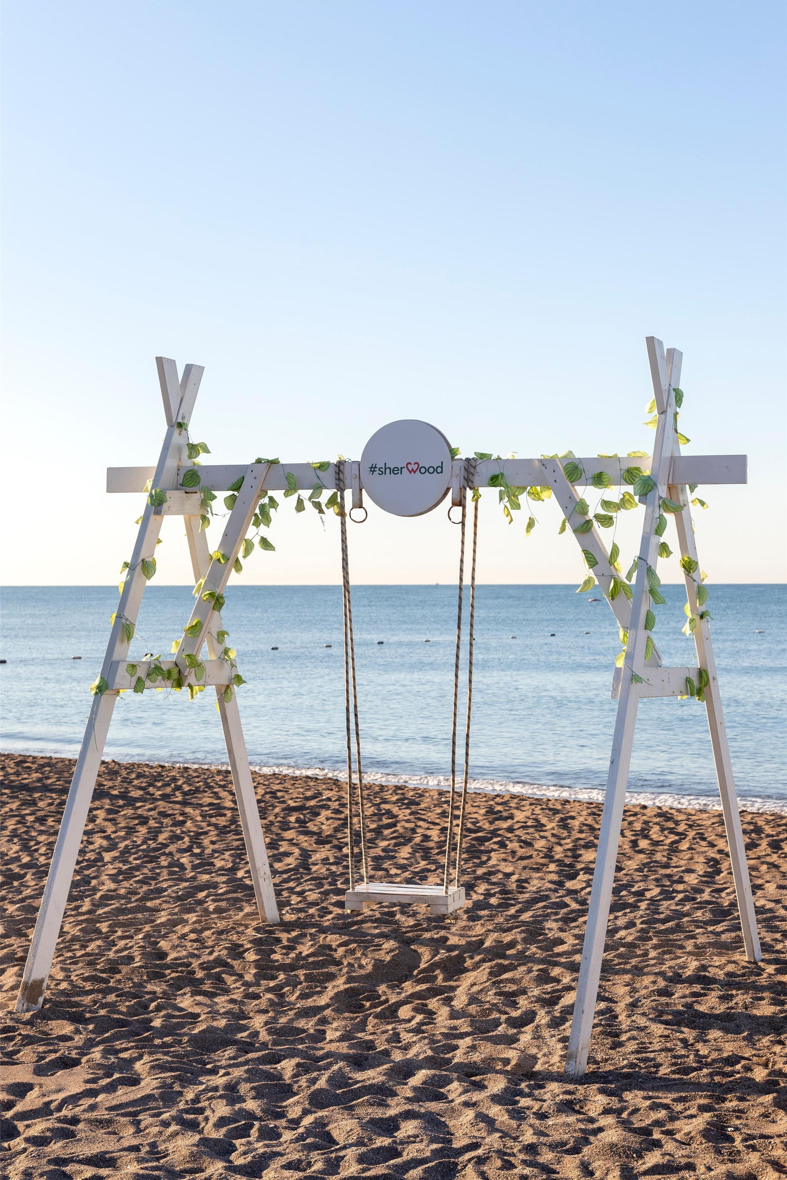 A beach swing set with a sign that reads "#sharewood" and is decorated with greenery.