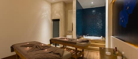 A modern spa room with a massage table, a wooden bench, a wooden counter with toiletries, and a blue-tiled wall.