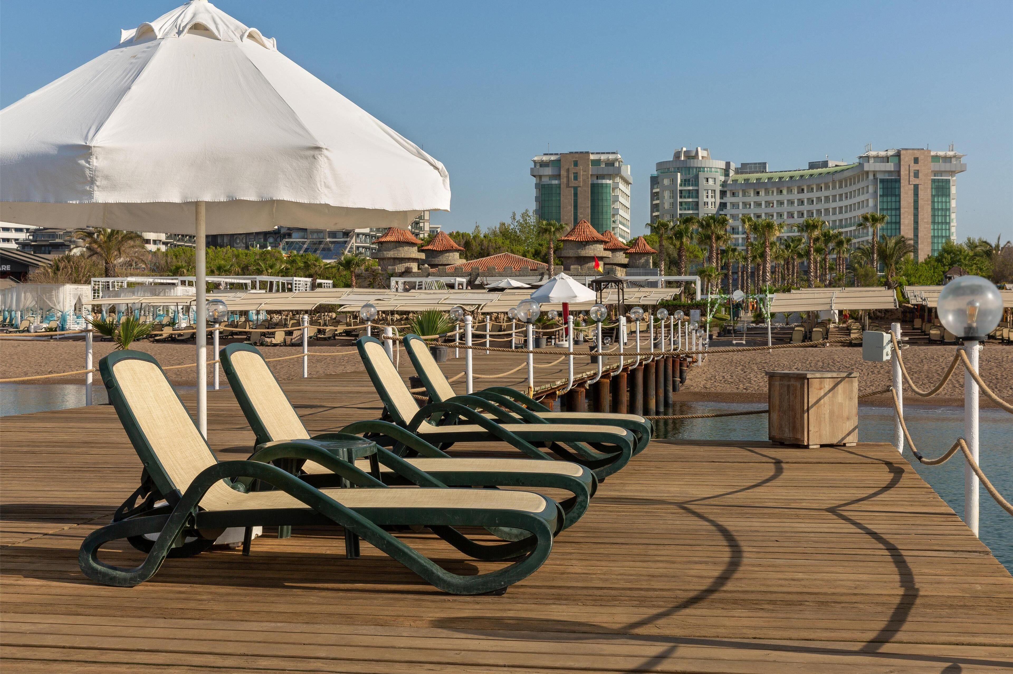 A wooden deck with lounge chairs and a large umbrella, overlooking a body of water with a view of hotel buildings in the background.