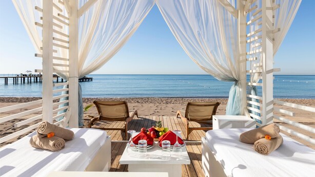 Een eetgedeelte aan het strand met uitzicht op de oceaan, met witte parasols, houten tafels en lounge stoelen.