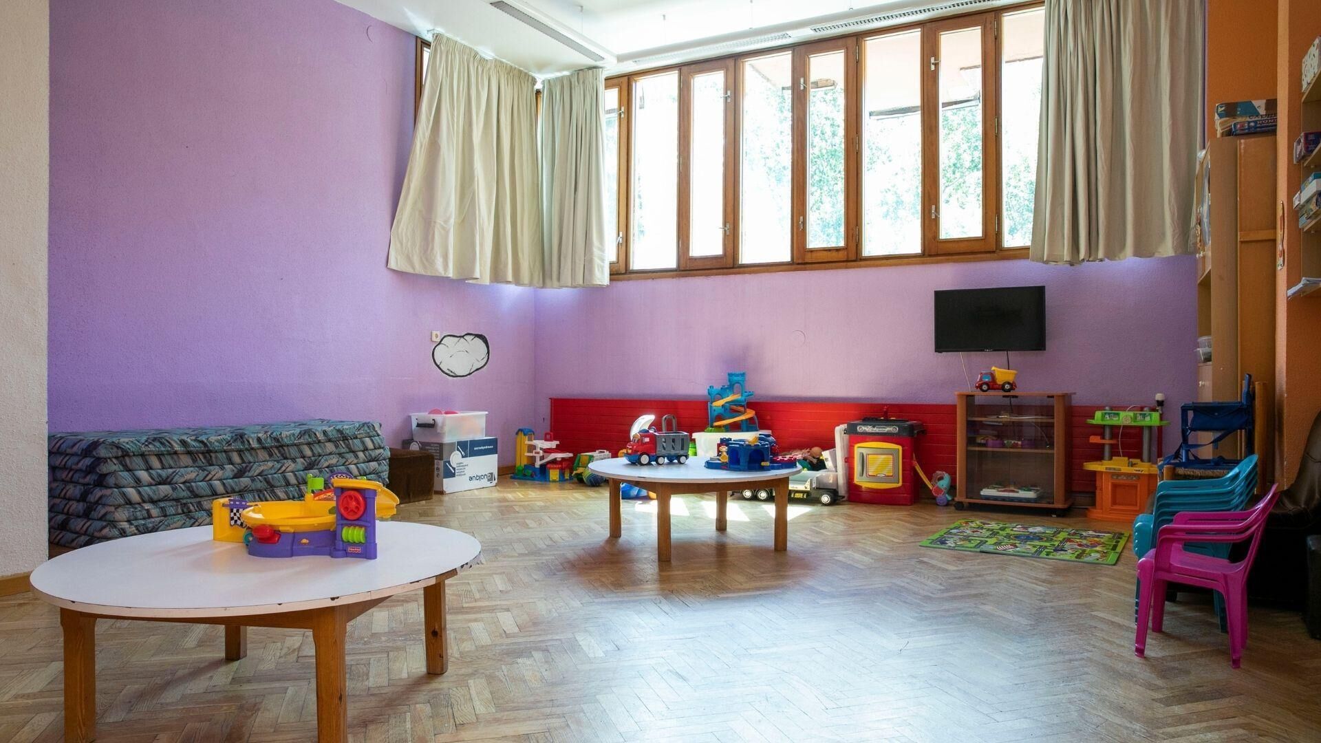 A room with a purple wall, a wooden floor, a round table, a sofa, a TV, and various toys.