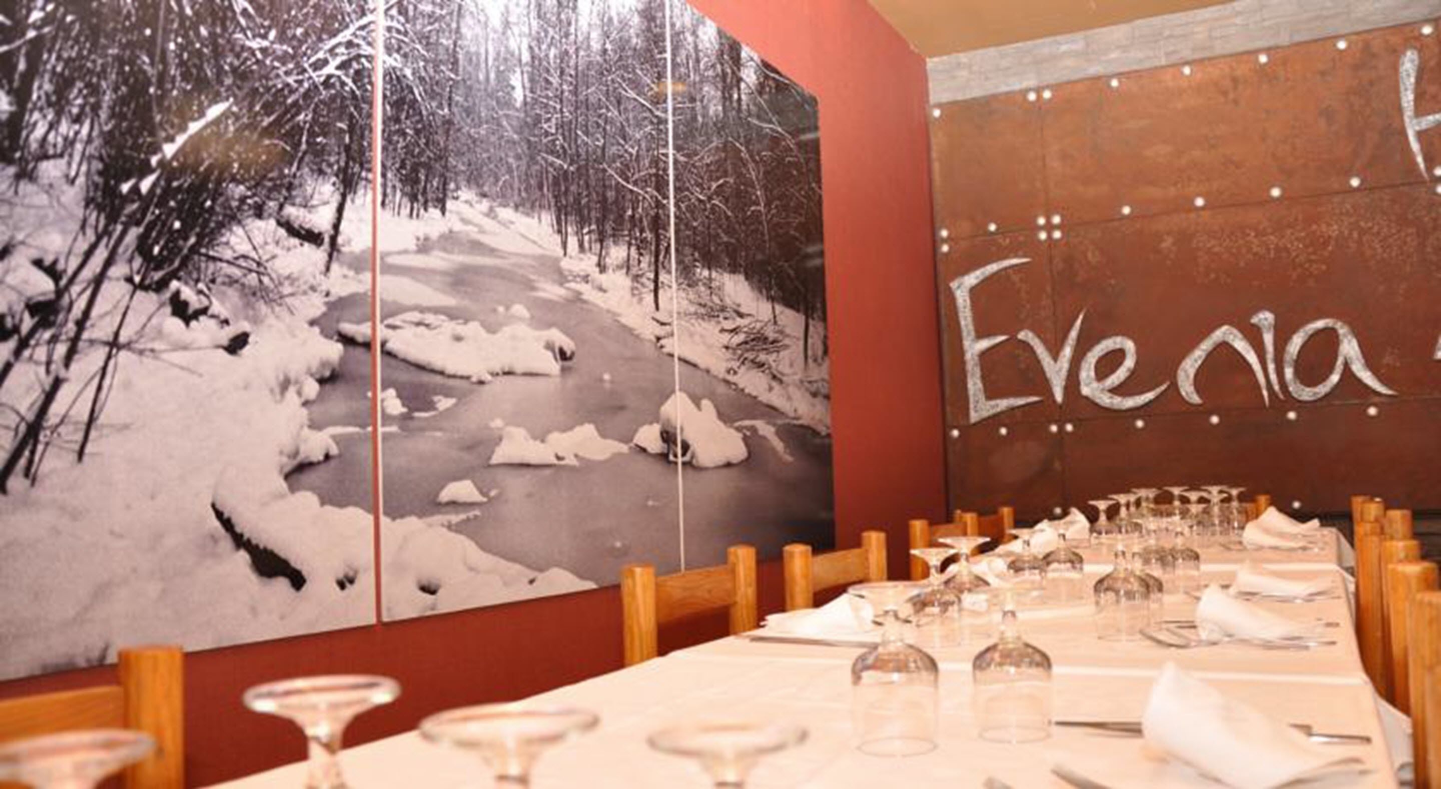 A restaurant with a long table set for dining, a wall with a snowy landscape painting, and the name "Evania" written on the wall.