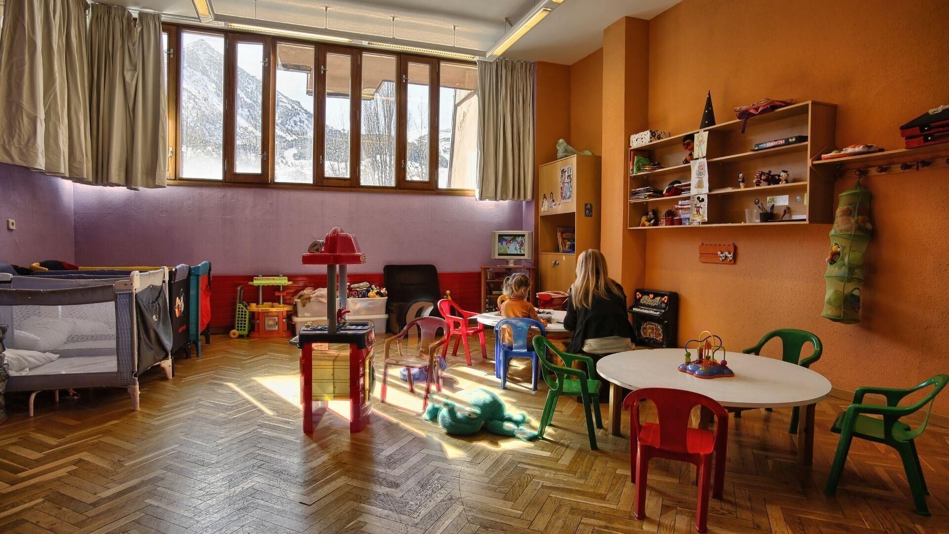 A children's playroom with a herringbone wooden floor, a playpen, a television, and a bookshelf filled with toys and books.