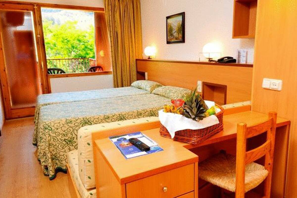 A hotel room with a bed, a desk with a basket of fruit, a chair, and a window with curtains.