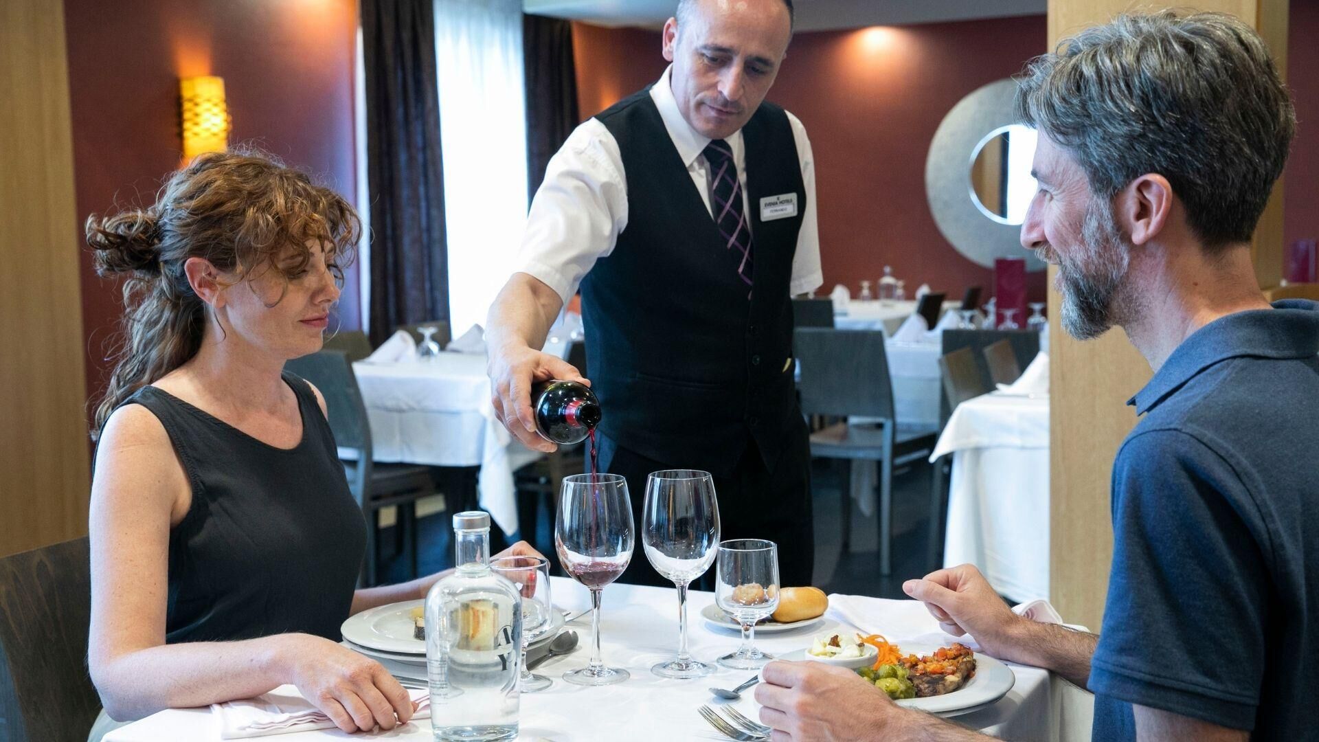 Ein Restaurantambiente mit einem Kellner, der Wein in Gläser einschenkt, einem Tisch mit Speisen und einer Wasserflasche.