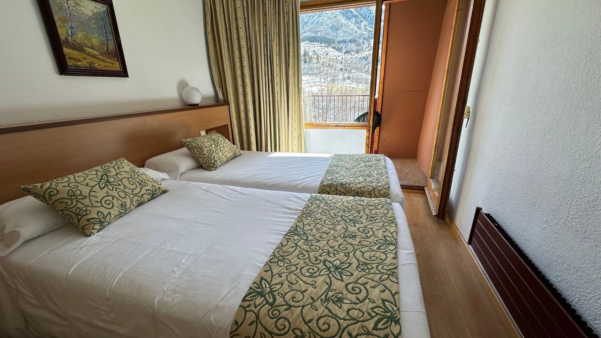 A hotel room with two beds, a painting on the wall, and a window with a view of snow-covered mountains.