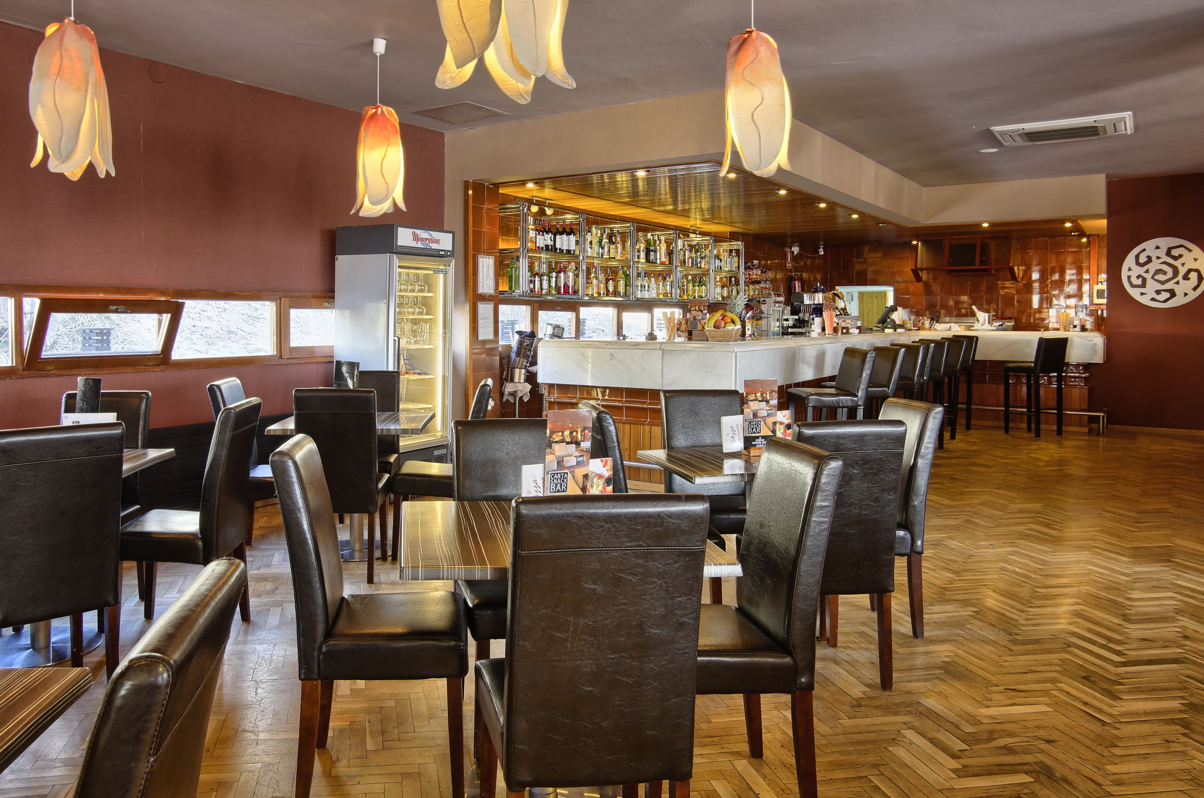 A restaurant with wooden flooring, leather chairs, a bar area with a variety of bottles, and a large decorative wall piece.