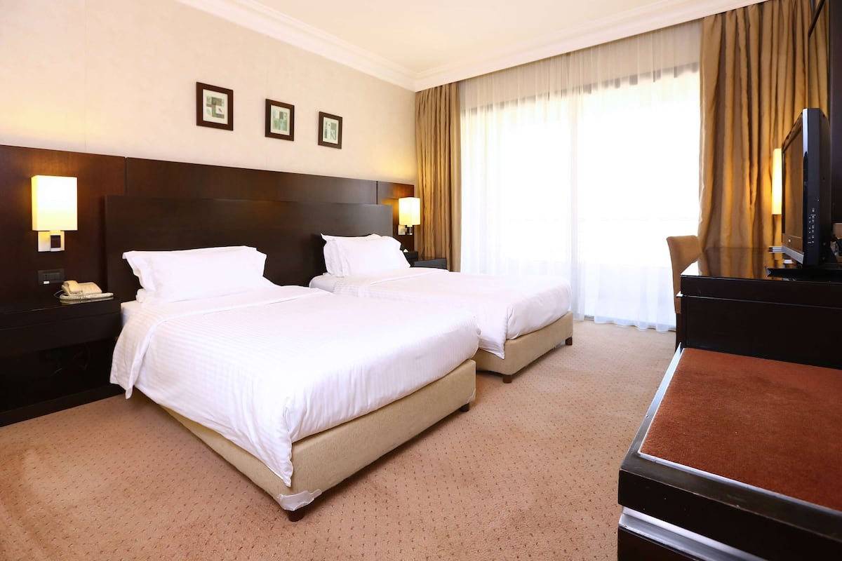 Ein Hotelzimmer mit zwei Betten, einem Schreibtisch mit Fernseher und einem großen Fenster mit Vorhängen.