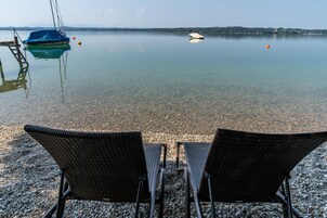 A clear lake with a dock and a boat, two wicker chairs facing the water, and a rocky shore.