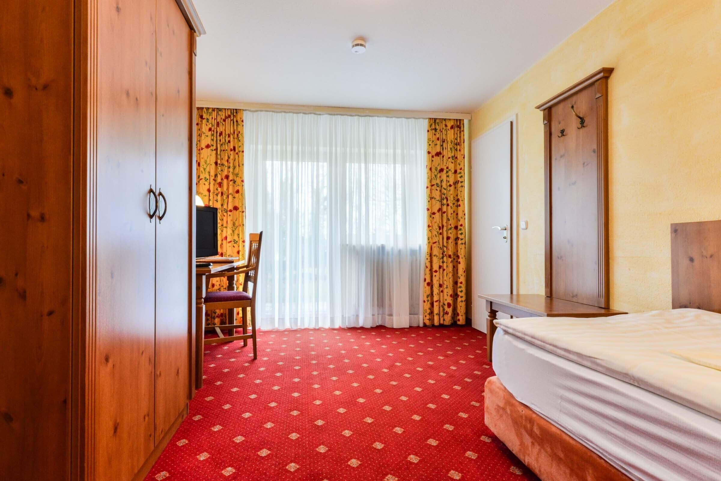 A hotel room with a red carpet, wooden wardrobe, a bed, a desk, and a chair.
